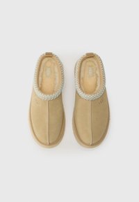 Tan suede slip-on shoes with a soft, cream-colored lining, featuring a decorative light blue stitching along the collar and a branded logo.