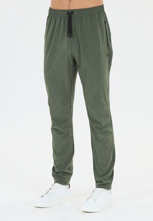 Mann trägt olivgrüne Hose mit Kordelzug und weiße Sneakers und steht vor einem schlichten weißen Hintergrund.