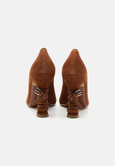 Καφέ ψηλοτάκουνα από σουέντ με μοναδικά σχεδιασμένες μαζεμένες φτέρνες που διαθέτουν πολλαπλούς ιμάντες και ασημένιες αγκράφες, που φαίνονται από πίσω.