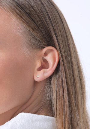 Goldener Stecker mit quadratischem Design, der einen einzelnen klaren Edelstein in der Mitte zeigt, vor einem glatten Hauthintergrund.