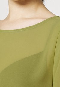 OLIVGRÜNE CHIFFONBLUSE MIT WEITEM HALSANSCHNITT UND WEICHER TEXTUR, MIT FALDENDRAPE UND UNAUFFÄLLIGEM SCHIMMER.