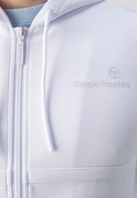 Sweat à capuche blanc avec fermeture éclair, fabriqué en matériau lisse et extensible. Présente un logo minimaliste sur la poitrine et des cordons ajustables sur la capuche.