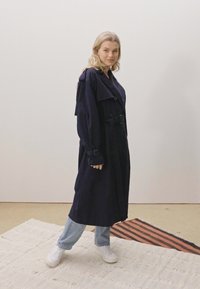 Cappotto trench navy con bottoni doppi, colletto ampio e cintura. Abbinato a jeans azzurri e sneakers bianche su un tappeto tessuto.