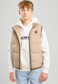 Beige värillinen toppaliivi mustalla reunuksella, vetoketjusuljenta ja kaksi etutaskua. Päällä valkoinen huppari, jossa tekstigrafiikkaa. Siniset farkut ja vaalea tausta.