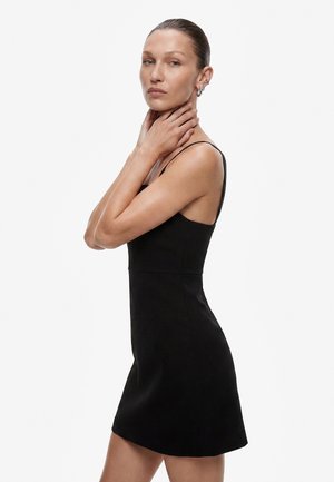 Woman with slicked-back hair wearing a sleeveless black dress, touching her neck with both hands, looking calmly ahead on a white background.