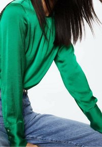 Grüne, satinierte Langarmbluse mit einem lockeren Schnitt, ausgestattet mit geknöpften Manschetten und einer glatten, glänzenden Textur. Kombiniert mit hochtaillierten Jeans.