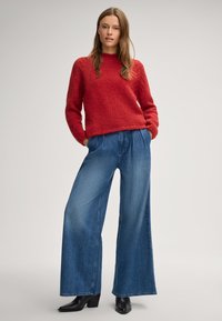 Maglione rosso lavorato a maglia con un motivo testurizzato e scollo a girocollo, abbinato a pantaloni in denim blu a gamba larga e stivaletti neri con tacco.