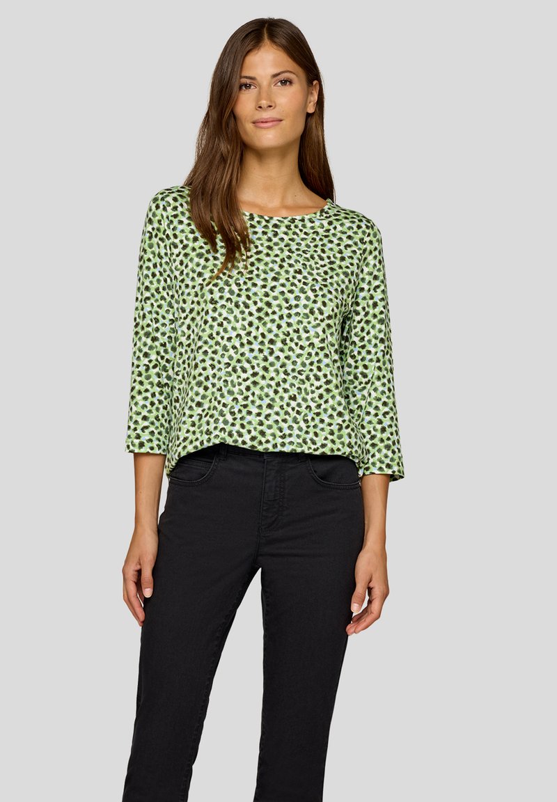 Grünes Baumwolloberteil mit rundem Ausschnitt, dreiviertel Ärmeln und einem schwarz-dunkelgrünen gepunkteten Muster. Kombiniert mit schwarzen Jeans.