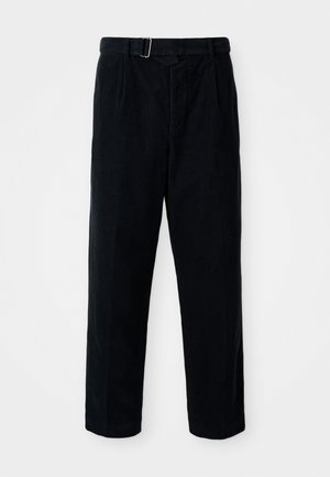 Grå hettegenser med rund hals og trykt etikett, kombinert med marineblå bukser og brune sneakers med kremfargede lisser og teksturert såle.