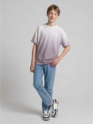 Garçon debout, une main sur la hanche, portant un t-shirt violet et blanc délavé, un jean bleu clair et des baskets blanches avec des accents foncés.