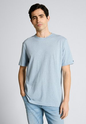 Mężczyzna o ciemnych włosach, ubrany w jasnoniebieską koszulę z krótkim rękawem i jasnoniebieskie dżinsy, stojący z jedną ręką w kieszeni na szarym tle.