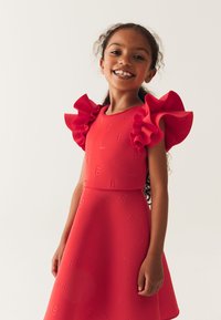 Robe rouge avec des manches à volants, texturée avec des motifs en "B" et des nœuds. Tissu lisse, corsage ajusté et jupe évasée.