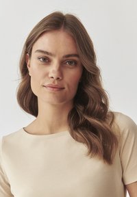 Kreemi värvi topp, millel on ümar kaelus, lühikesed varrukad ja sile tekstuur; mudelil on laineline pruun juuksed ja loomulik meik.