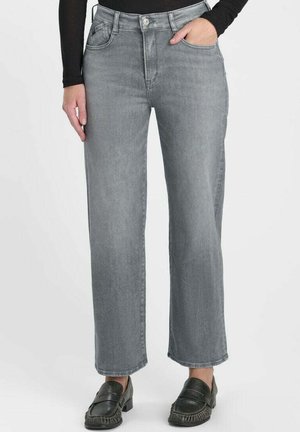 Persoon draagt grijze jeans met hoge taille en rechte pijpen, zwarte loafers en een zwart shirt met lange mouwen, staand met één hand in de broekzak.
