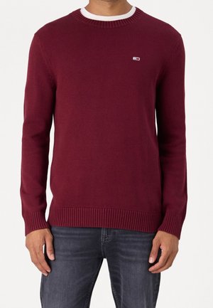 Homme portant un pull en maille bordeaux ajusté avec des poignets et un bas côtelés et un petit logo sur la poitrine gauche, associé à un jean bleu foncé.