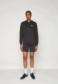 Czarna rozpinana bluza z kapturem z przednią kieszenią i logo, połączona z ciemnymi szortami. Model nosi białe skarpetki i czarne buty sportowe.