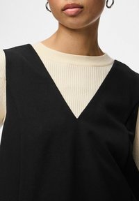 Černé šaty s výstřihem do V vrstvené přes krémový žebrovaný top s dlouhými rukávy. Disponují hladkou texturou a kontrastními barvami, jsou viditelné kruhové náušnice.
