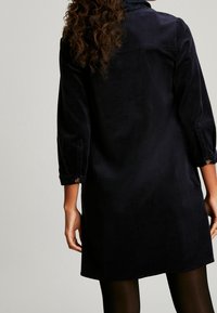 Granatowa sukienka z korduroyu z długimi rękawami, zapięciem na guziki i kołnierzykiem. Rozkloszowany fason i gładka tekstura z prostym, eleganckim wzorem.