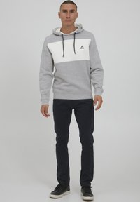 Grijze hoodie met een witte horizontale streep, voorzien van een driehoekig logo, gecombineerd met donkere broek en zwarte sneakers.