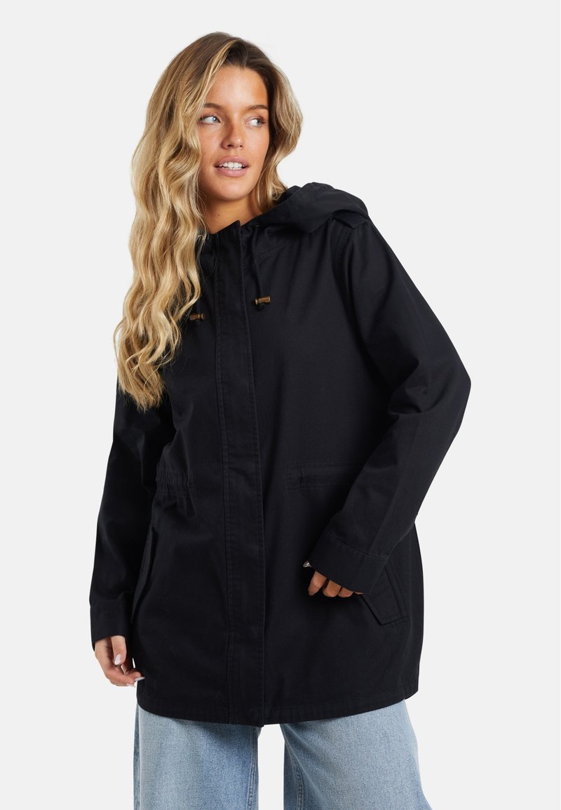 Veste à capuche noire en tissu lisse, avec des poches latérales, un cordon de serrage réglable et une coupe décontractée. Portée sur un jean bleu clair.