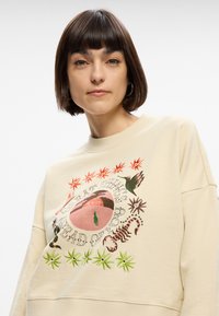 Beige mikina s kulatým výstřihem a volným střihem. Centrální grafika obsahuje barevné vyšívání flóry a fauny. Směs bavlny.
