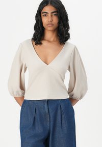 Top texturado bege com decote em V, mangas bufantes e cintura ajustada. Combinado com calças azuis de cintura alta com pregas.