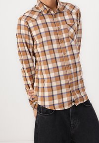 Man in een bruine, witte en blauwe geruite button-up shirt met zwarte jeans, hand op zijn onderrug, staand tegen een witte muur.