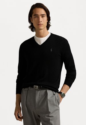 Pull noir à col en V en maille fine, associé à un pantalon gris plissé. Logo notable sur la poitrine gauche. Le modèle a les cheveux de longueur moyenne.