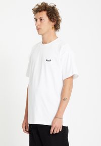 Junger Mann mit lockigem Haar, der ein weißes Volcom-T-Shirt, schwarze Hose, orange Pendelohringe, ein Stern-Tattoo am Unterarm und ein silbernes Armband trägt.