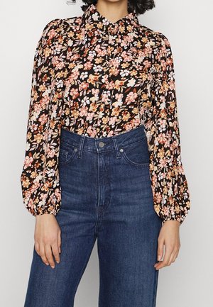 Schwarze Bluse mit floralem Muster in Pink und Orange, hohem Kragen und Puffärmel. Kombiniert mit dunkelblauen, hochgeschnittenen Jeans.