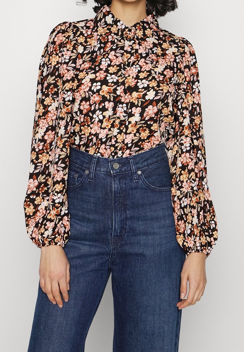 Blusa nera floreale con fiori rosa e arancioni, collo alto e maniche a sbuffo. Abbinata a jeans a vita alta blu scuro.