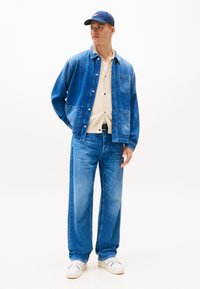 Denim jacket in medium blue with two pockets, paired with light blue wide-leg jeans. Underneath, a cream knit shirt with buttons. White sneakers.