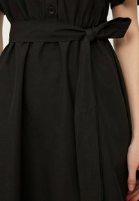 Robe noire avec une taille cintrée, dotée d'une ceinture en tissu nouée. Le matériau est lisse, avec des manches courtes et des détails de boutons à l'avant.