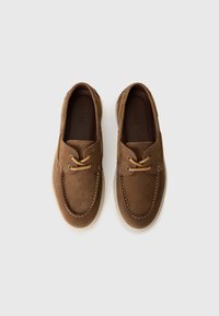 Een paar bruine suède bootschoenen met tan kleurige veters en witte rubberen zolen, van bovenaf bekeken tegen een witte achtergrond.