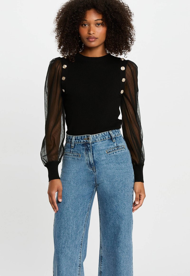 Frau mit lockigem Haar, die ein schwarzes Oberteil mit transparenten Puffärmeln und goldenen Knöpfen trägt, kombiniert mit hoch taillierten, weit geschnittenen blauen Jeans.