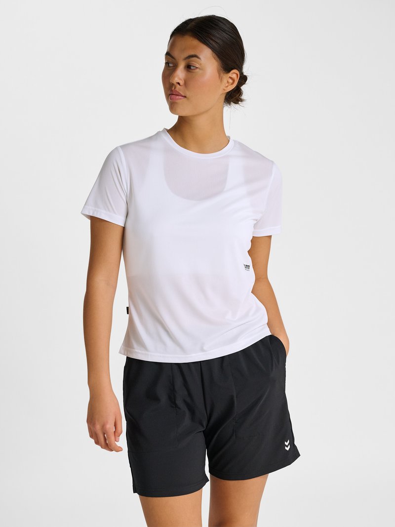 Frau trägt ein weißes kurzärmeliges Oberteil und schwarze Sportshorts, hat die Hände in den Taschen und schaut nach links vor weißem Hintergrund.