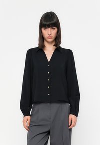 Blusa preta com decote em V, mangas longas e detalhes em botões dourados. Tecido leve com uma superfície ligeiramente texturada. Combinada com calças cinzas.