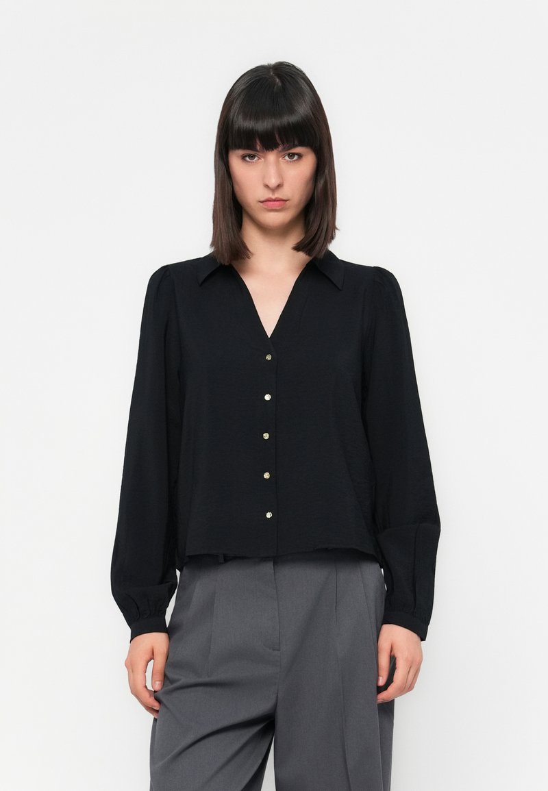 Blusa preta com decote em V, mangas longas e detalhes em botões dourados. Tecido leve com uma superfície ligeiramente texturada. Combinada com calças cinzas.