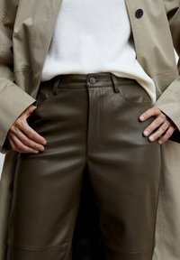 Braune Lederhose mit glatter Textur, ausgestattet mit Vordertaschen und einem Metallknopfverschluss, kombiniert mit einem weißen Pullover und einem beigefarbenen Mantel.