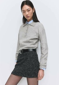 Pulover gri cu mâneci lungi, cu guler și detalii cu nasturi, asortat cu o fustă neagră texturată și o curea neagră lucioasă. Manșetele albastre ale cămășii sunt vizibile.