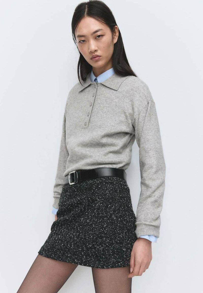 Pulover gri cu mâneci lungi, cu guler și detalii cu nasturi, asortat cu o fustă neagră texturată și o curea neagră lucioasă. Manșetele albastre ale cămășii sunt vizibile.