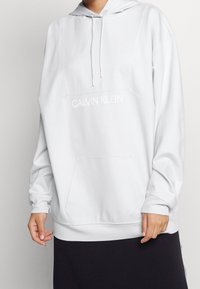 Personne portant un sweat à capuche blanc de Calvin Klein avec une poche avant et des manches longues, associé à un bas noir.