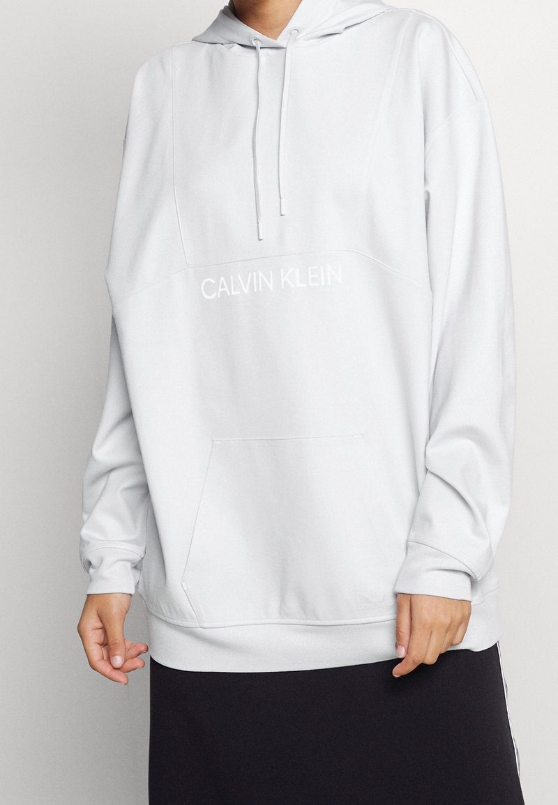 Personne portant un sweat à capuche blanc de Calvin Klein avec une poche avant et des manches longues, associé à un bas noir.