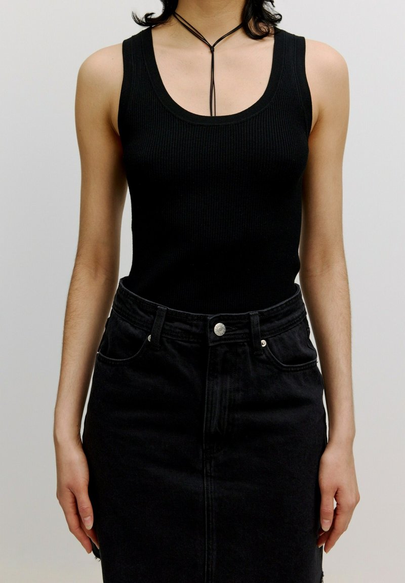 Schwarzes geripptes Tanktop kombiniert mit einem hochtaillierten schwarzen Jeansrock. Verfügt über eine glatte Textur und sichtbare Knopfdetails.
