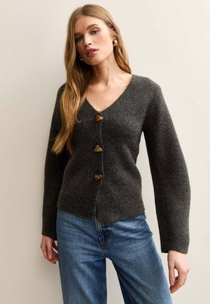 Mujer con cárdigan gris oscuro de cuello en V con grandes botones en forma de triángulo y jeans azules, de pie frente a una pared lisa y clara.