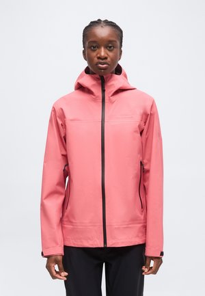 Donna che indossa una giacca impermeabile rosa con cappuccio, zip nera e tasche laterali con zip, in piedi davanti a uno sfondo chiaro e uniforme.