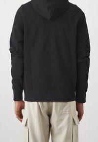 Sudadera negra con capucha, de manga larga y con ribete en el dobladillo. Lleva puesta sobre pantalones cargo de color claro, con detalles de bolsillos laterales.