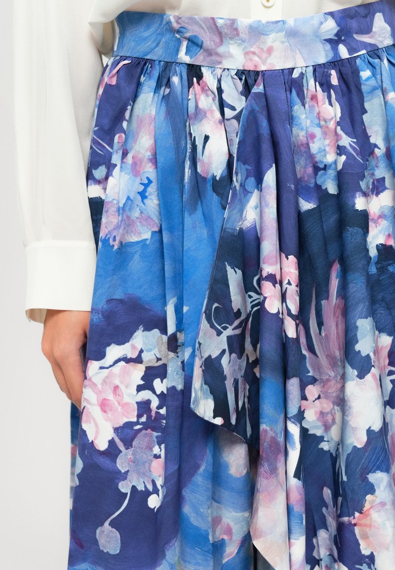 Donna che indossa una gonna floreale blu e viola con tessuto a strati e una blusa bianca a maniche lunghe, con una mano appoggiata al suo fianco.