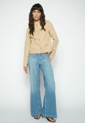 Žena v béžovom zapínanom saku a širokých modrých džínsach s vzorovanými pásmi, stojaca pred jednoduchým bielym pozadím.