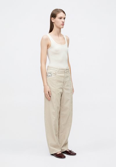 Μπεζ παντελόνι φαρδιάς γραμμής με υφή υφάσματος και τσέπη με σχέδιο, συνδυασμένο με εφαρμοστό κρεμ τοπ χωρίς μανίκια και λαμπερά μπορντό παπούτσια.